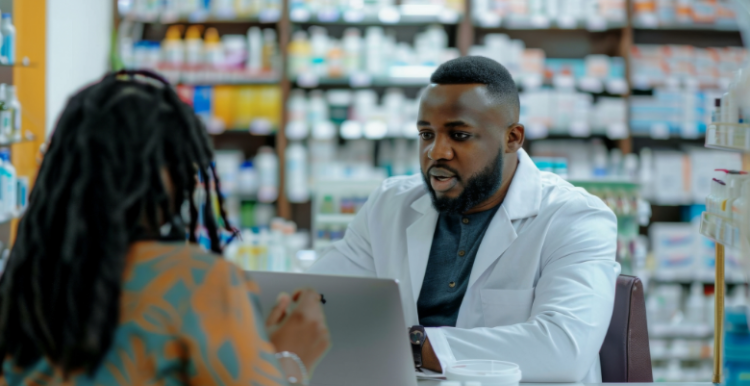 A pharmacist is looking at their laptop whilst a person is sat across the countr from them. Behind them are rows of medication packages