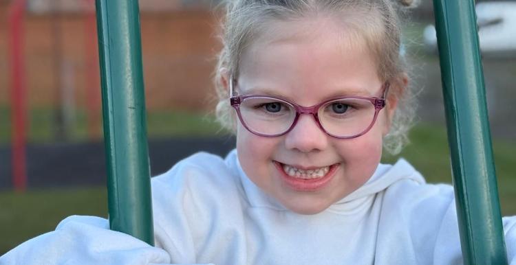 A young girl is leaning on a climbing frame in an outside playground. She is smiling at the camera and is wearing glasses.