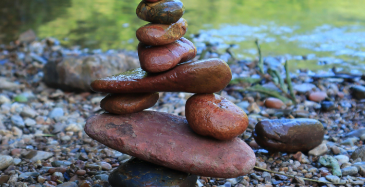 Photo of some rocks balancing on top and beside other rocks and flat stones. The rocks are beside some water.