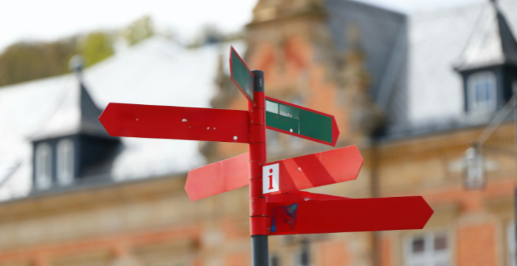 A signpost can be seen with nothing on it other than an 'i' for information. A building is blurred in the background.