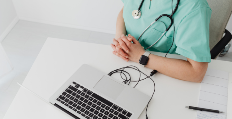 A person with a stethoscope hanging round thier neck is sat beside a laptop computer.