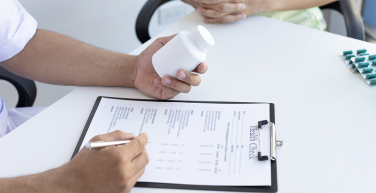 Image of a table - someone is looking at a bottle of pills and writing something on a clipboard. Another person can just be seen to the side, sitting with their hands in their lap.