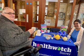 Photo of 2 people sat either side of a table which has the Healthwatch Surrey tablecloth on it. The person in the forground of the image is sat in a wheelchair. They are both having a cup of tea.