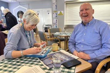 Photo of one of our engagment team sitting at a table taking notes whilst a man sat next to her is smiling at the camera.