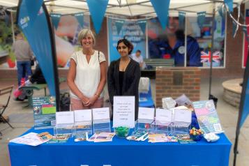 A photo of 2 of our volunteers stood outside. They are under a gazebo which has Healthwatch Surrey bunting around it. They are stood behind table that is covered with a Healthwatch Surrey branded tablecloth. There is lots of leaflets and merchandise on the table.