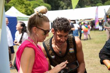 Photo of an outside event with tents in the background, and people can be seen with rainbow flags. In the foreground 2 people are stood looking at something. On eof the people has a rainbow badge on, the other is in a bright pink top.