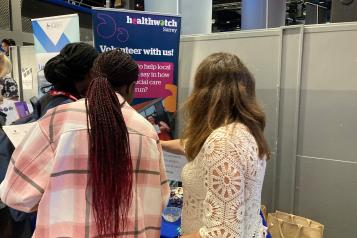 Photo of our volunteer officer talking to 2 students. In the background is our Healthwatch Surrey 'Volunteer with us' banner