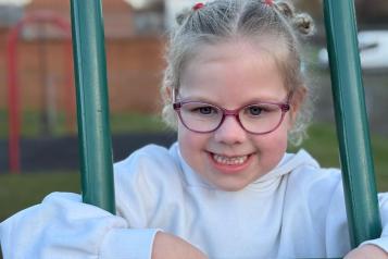 A young girl is leaning on a climbing frame in an outside playground. She is smiling at the camera and is wearing glasses.