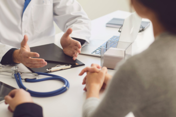 A close up of 2 people sitting opposite each other - the photo mainly shows their hands and forearms resting on the table. On the table is also a clipboard and a stethoscope.