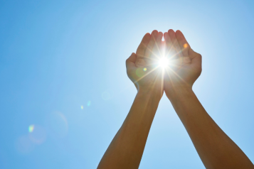 Two hands are raised towards the sun with the sunlight shining between the 2 open palms.