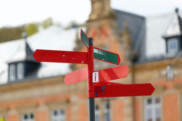 A signpost can be seen with nothing on it other than an 'i' for information. A building is blurred in the background.
