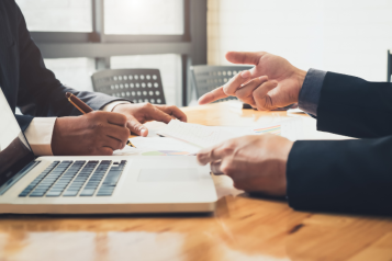 The hands of 2 people can be seen on a table. One is pointing to what the other person is writing down. In the foreground is a laptop computer.