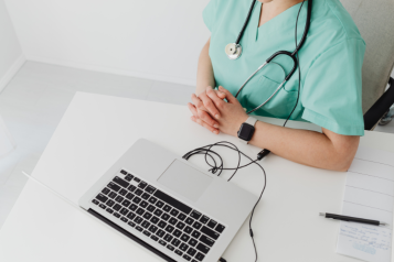 A person with a stethoscope hanging round thier neck is sat beside a laptop computer.
