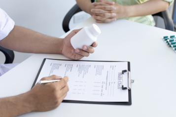 Image of a table - someone is looking at a bottle of pills and writing something on a clipboard. Another person can just be seen to the side, sitting with their hands in their lap.