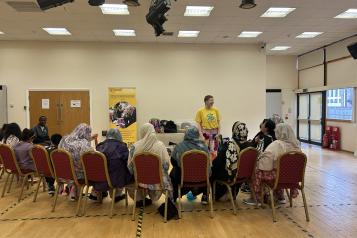 Photo of a person with a tambourine is stood facing a group of women who are sat in chairs facing away from the camera. The  person with the tambourine is wearing brightly coloured clothes, the women sitting down are all wearing headscarves. 