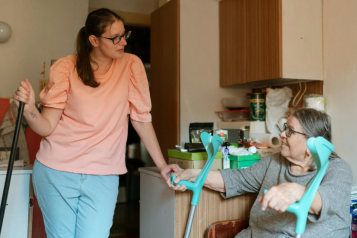 Photo of inside someone's home. One person is sitting down holding elbow style crutches. Another person is standing up holding something in her right hand (a broom or mop handle?) and leaning on the counter. They are talking to each other.