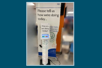 Photo of a hospital pull up banner which says 'Please tell s how we're doing today. In front of it is a screen where you can provide feedback - it is showing a row of circles with facila expreseesions on them - coloured in a range from green to red.