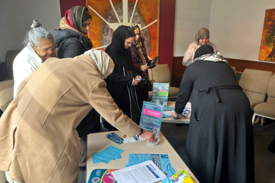 A group of women wearing head scarves are looking at information laid out on a table. There is a Healthwatch Surrey leaflet displayed on the table.