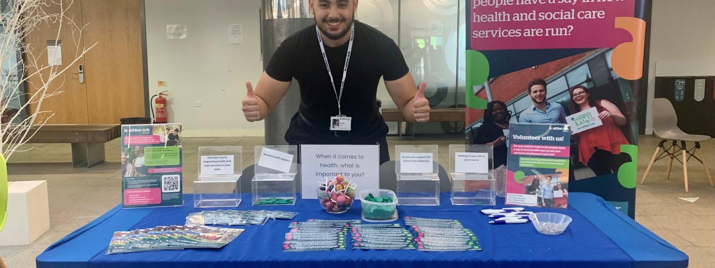 One of our volunteers is standing behind a table and making thumbs up signs at the camera. The table has a Healthwatch Surrey table cloth on it and has merchandise and leaflets laid out on top.