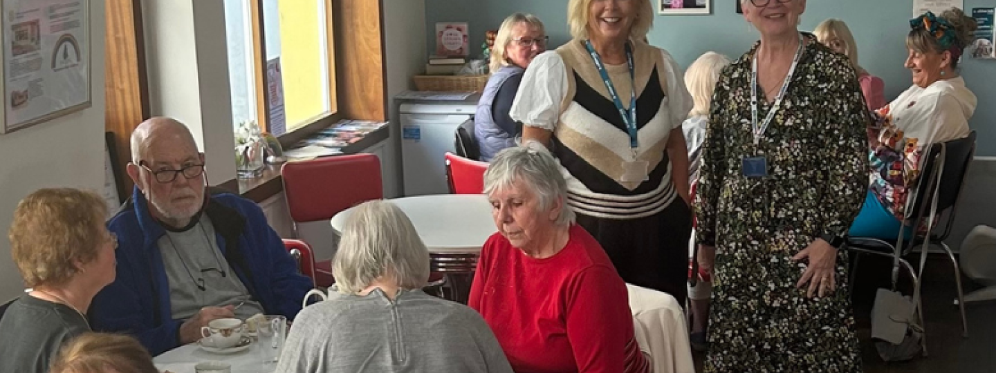 Two of our Insight and Engagement Officers at an Age Concern coffee morning. In the foreground are people sat around circular tables.