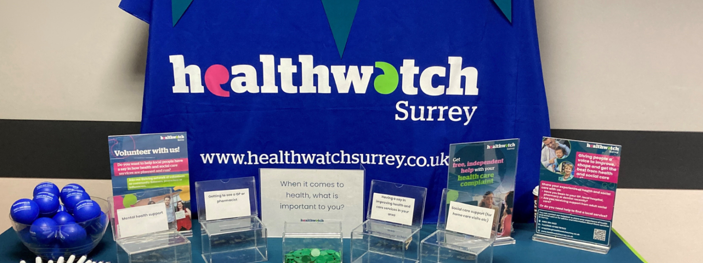 Image of a table covered with a blue tablecloth, with a Healthwatch Surrey sign behind it. The table has posters and some information on it.