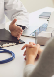 A close up of 2 people sitting opposite each other - the photo mainly shows their hands and forearms resting on the table. On the table is also a clipboard and a stethoscope.