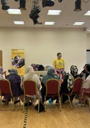 Photo of a person with a tambourine is stood facing a group of women who are sat in chairs facing away from the camera. The  person with the tambourine is wearing brightly coloured clothes, the women sitting down are all wearing headscarves. 