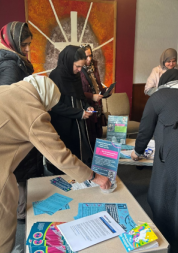 A group of women wearing head scarves are looking at information laid out on a table. There is a Healthwatch Surrey leaflet displayed on the table.