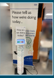 Photo of a hospital pull up banner which says 'Please tell s how we're doing today. In front of it is a screen where you can provide feedback - it is showing a row of circles with facila expreseesions on them - coloured in a range from green to red.