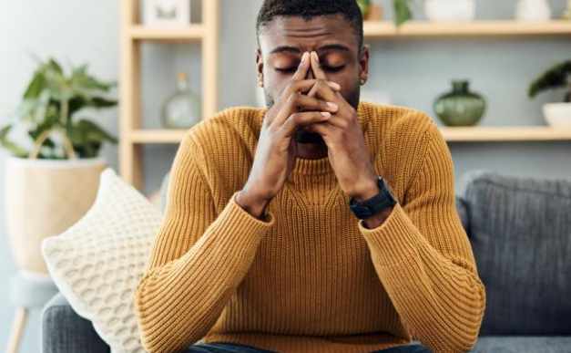 Man sat on a sofa. His elbows are resting on his knees and his hands are linked together in front of his face and his eyes are closed.