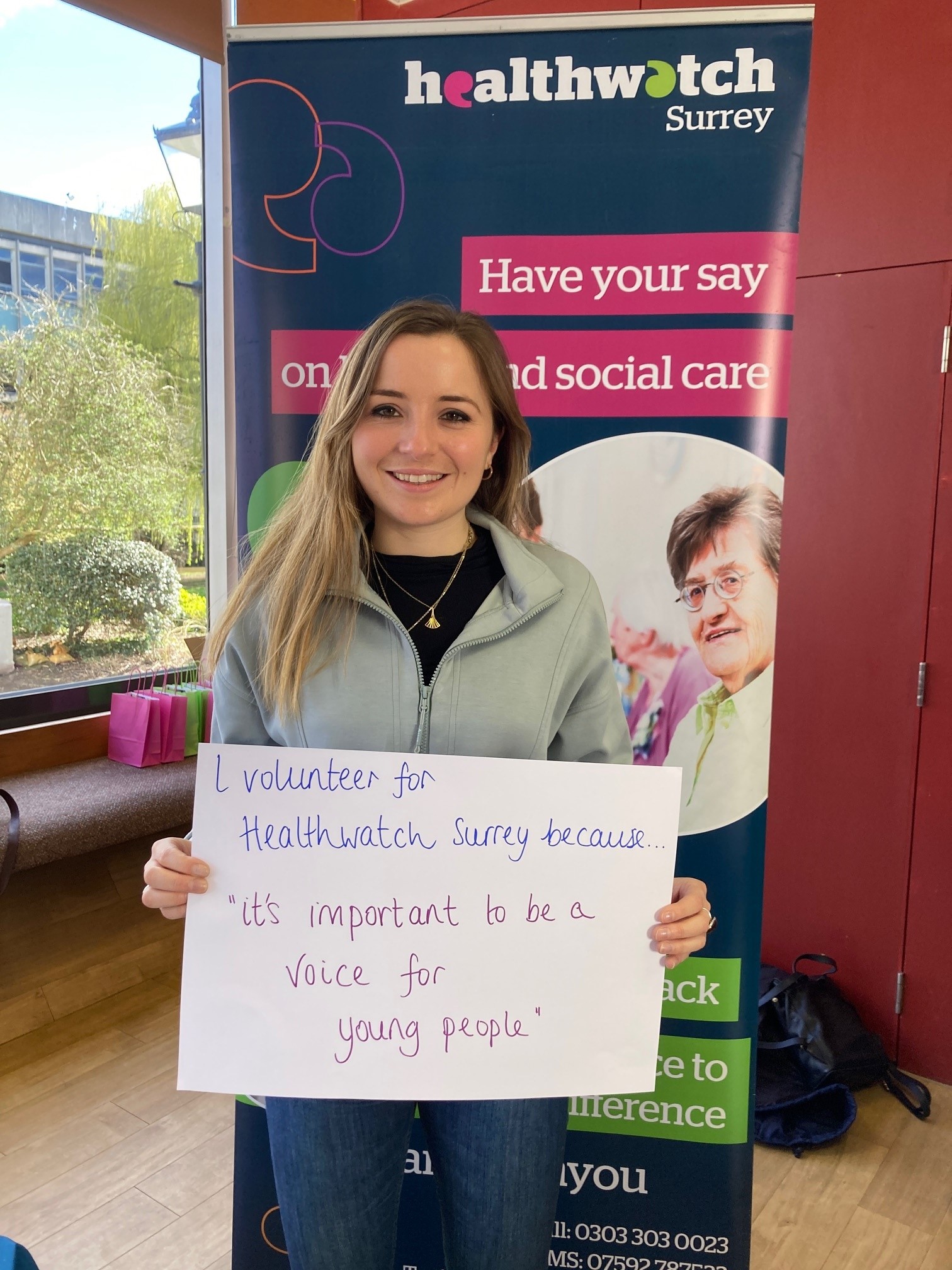 katie is stood in front of the Healthwatch Surrey banner and holds a card that reads 'I volunteer for Healthwatch Surrey because it's important to be a voice for young people.