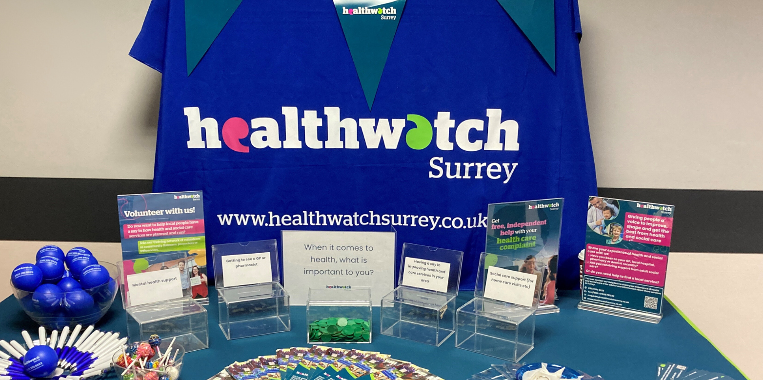 Photo of a table covered with a Healthwatch Surrey tablecloth, with Healthwatch Surrey bunting behind it. The table is laid out with leaflets, merchandise and some voting boxes asking, when it comes to health, what is important to you.